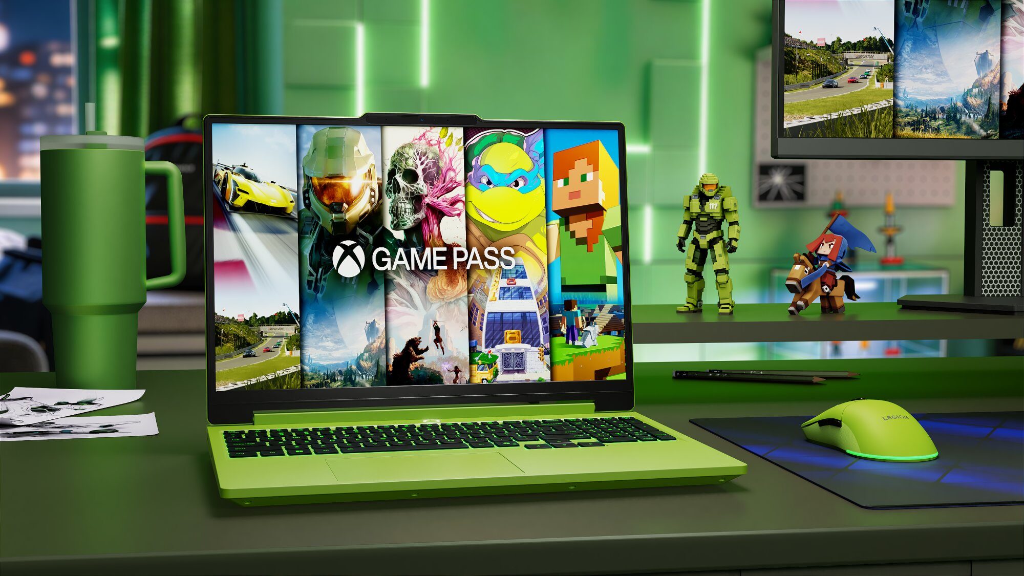 Zielony laptop z otwartą klapą, na ekranie którego wyświetlana jest grafika Xbox Game Pass z fragmentami gier, stoi na biurku obok zielonego kubka termicznego, myszki gamingowej i figurek postaci z gier.
