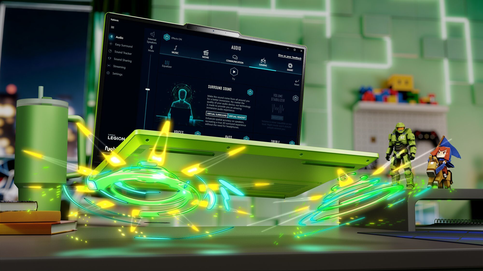 Zielony laptop Lenovo Legion otwarty na biurku, z włączonym oprogramowaniem Nahimic Audio, obok zielonego kubka termicznego, książek i figurek postaci z gry Halo oraz anime, w tle zielone, neonowe dekoracje.