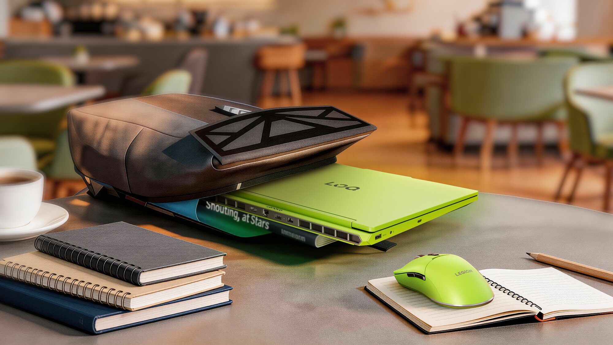Jasnozielony laptop marki Legion jest częściowo schowany pod ciemnobrązową torbą na stole w kawiarni, obok stosu notatników, filiżanki kawy i zielonej myszki komputerowej.
